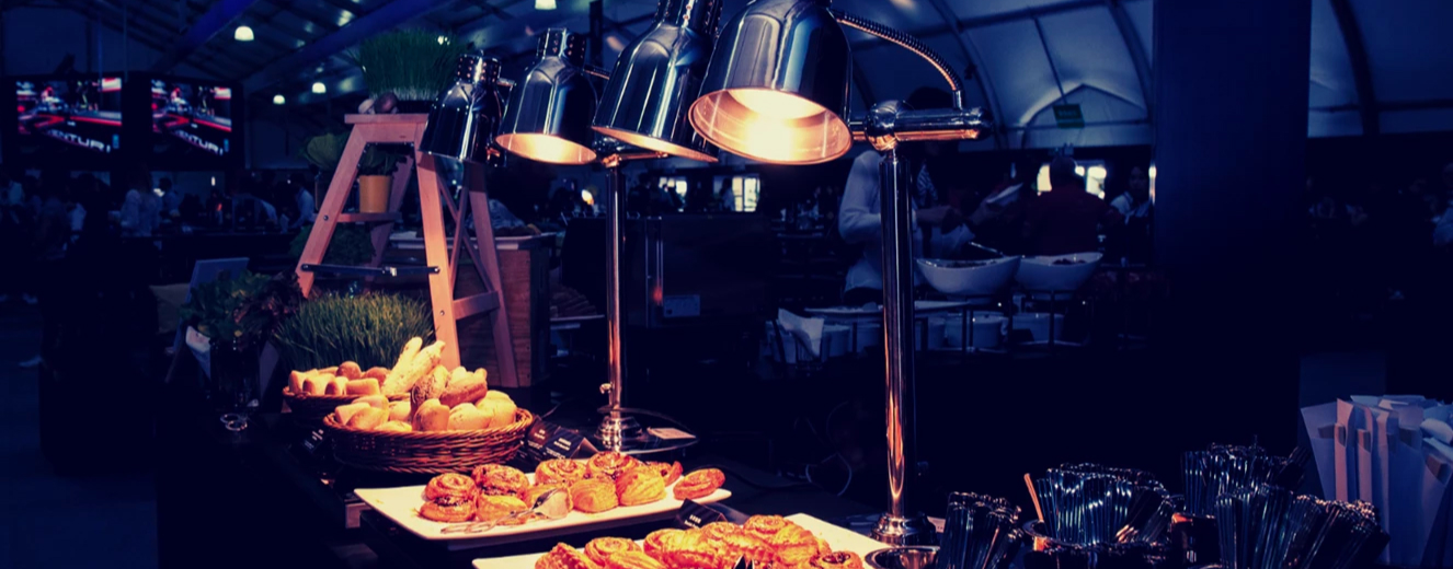 Warm-toned lamps illuminate a variety of pastries on a display table in a bustling setting.
