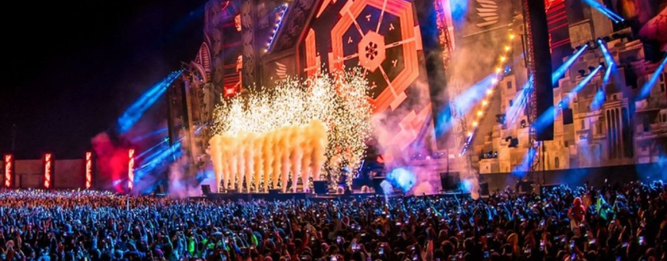 Vibrant stage with colourful lights and fireworks, surrounded by a large, cheering crowd at a festival.