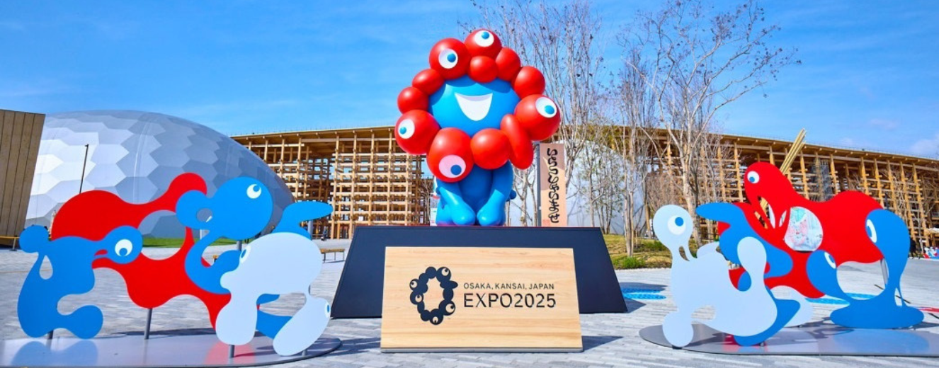 Bright blue mascot with red accents stands beside a wooden sign for EXPO 2025.