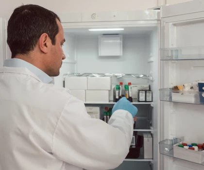 A man in a white lab coat retrieves a vial from a medical refrigerator with blue gloves.