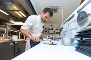 A chef in a white uniform carefully arranges food on a plate in a modern kitchen.