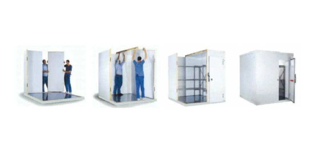 Two workers in blue uniforms assemble a modular cold room with white panels and shelves.