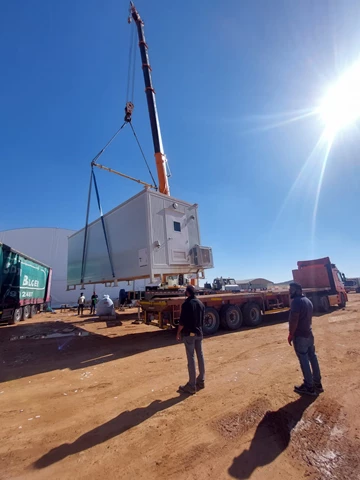 A crane lifts a large container against a clear blue sky, with two workers observing nearby.