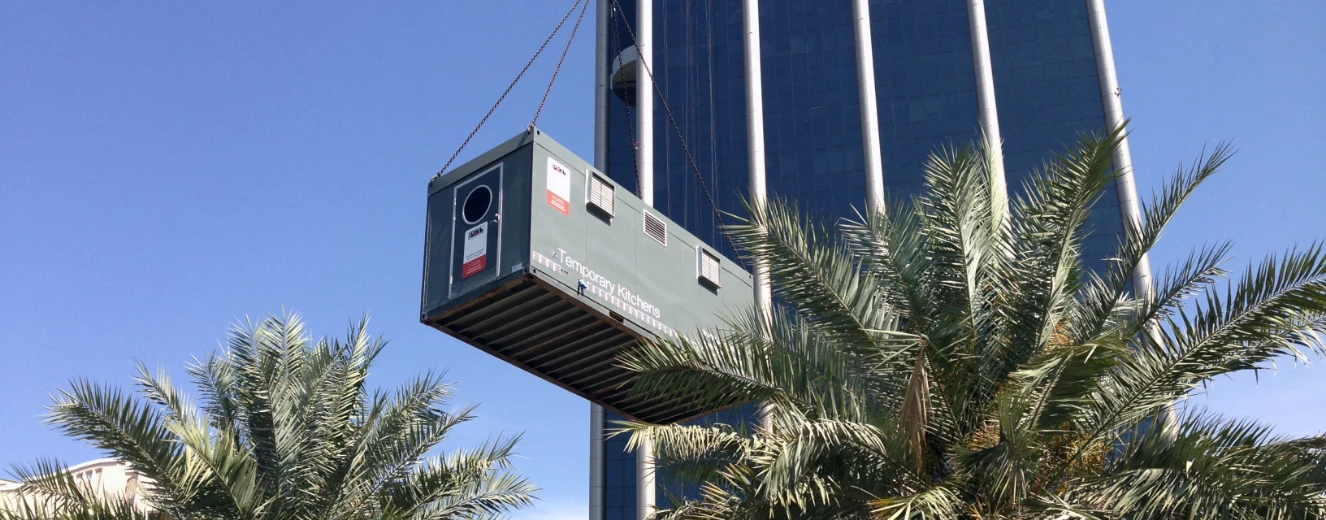 A green temporary kitchen container is suspended from a crane near a tall building and palm trees.