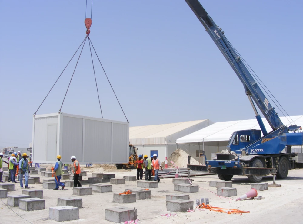 A crane lifts a large white container above grey concrete blocks on a construction site.