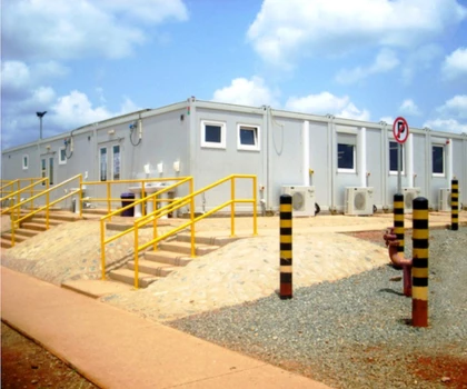 A white modular building with yellow railings, surrounded by sandy terrain and blue sky.