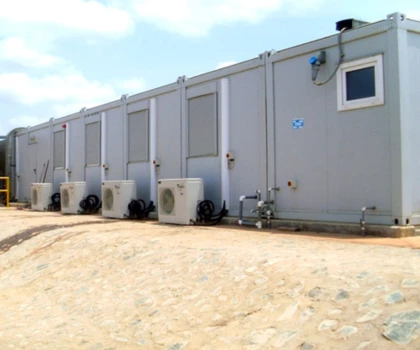 Grey modular units with air conditioning units, set against a sandy ground and blue sky.