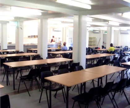 Large commercial kitchen space with wooden tables and black chairs, featuring white walls and bright lighting.