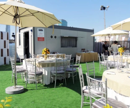 Temporary kitchen unit surrounded by white tables and chairs on green grass under large umbrellas.