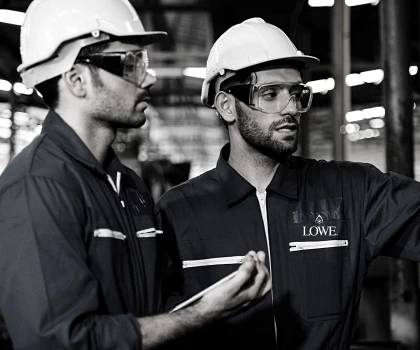 Two men in dark blue overalls and white helmets discuss a project in a workshop.