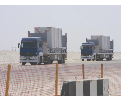 Three blue trucks transporting cargo on a sandy road, with a barrier visible in the foreground.