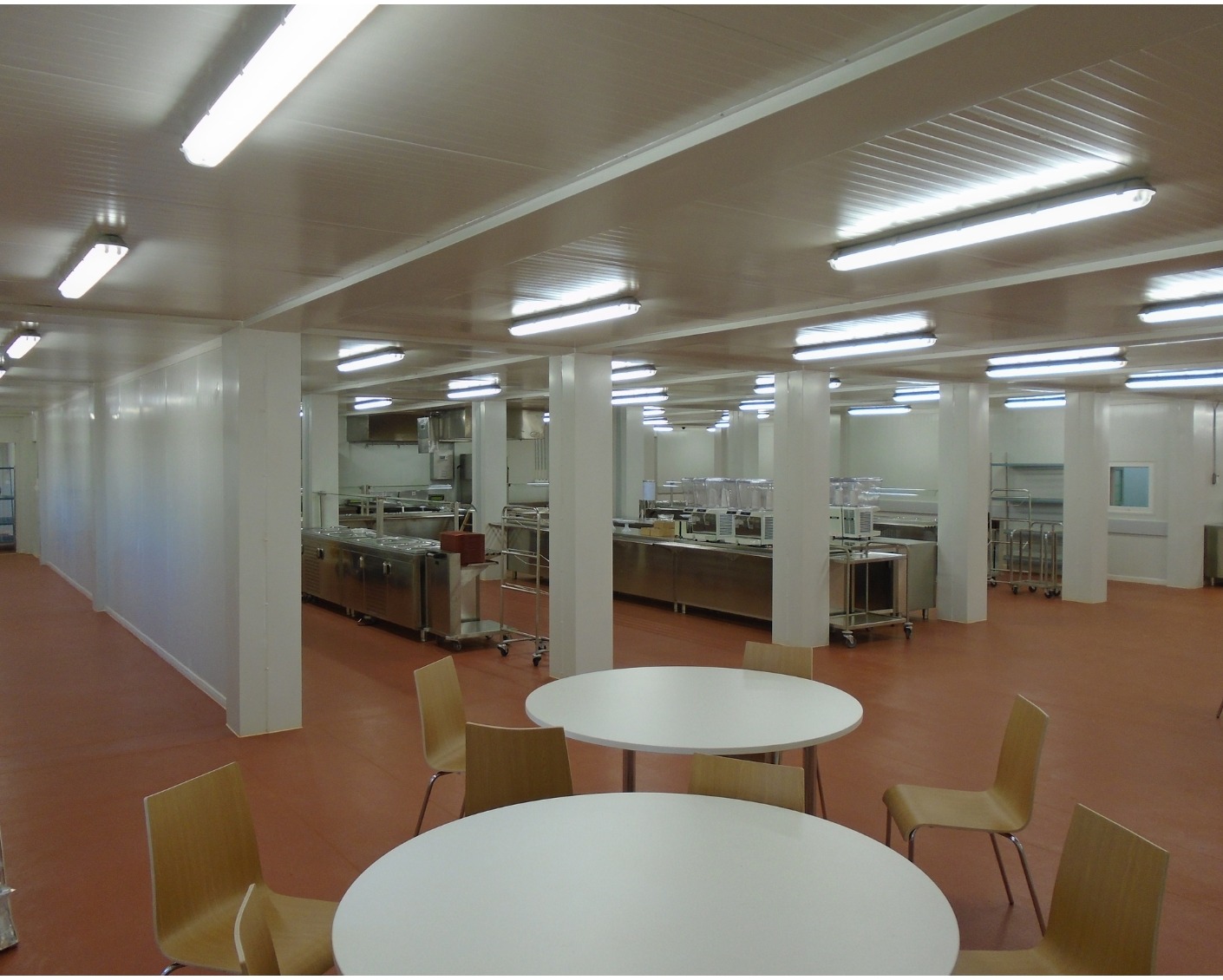 Brightly lit canteen featuring white tables, wooden chairs, and stainless steel catering infrastructure in the background.