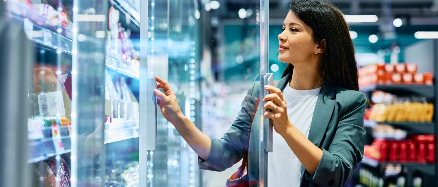 A woman in a grey jacket examines products in a brightly lit refrigerated display case.