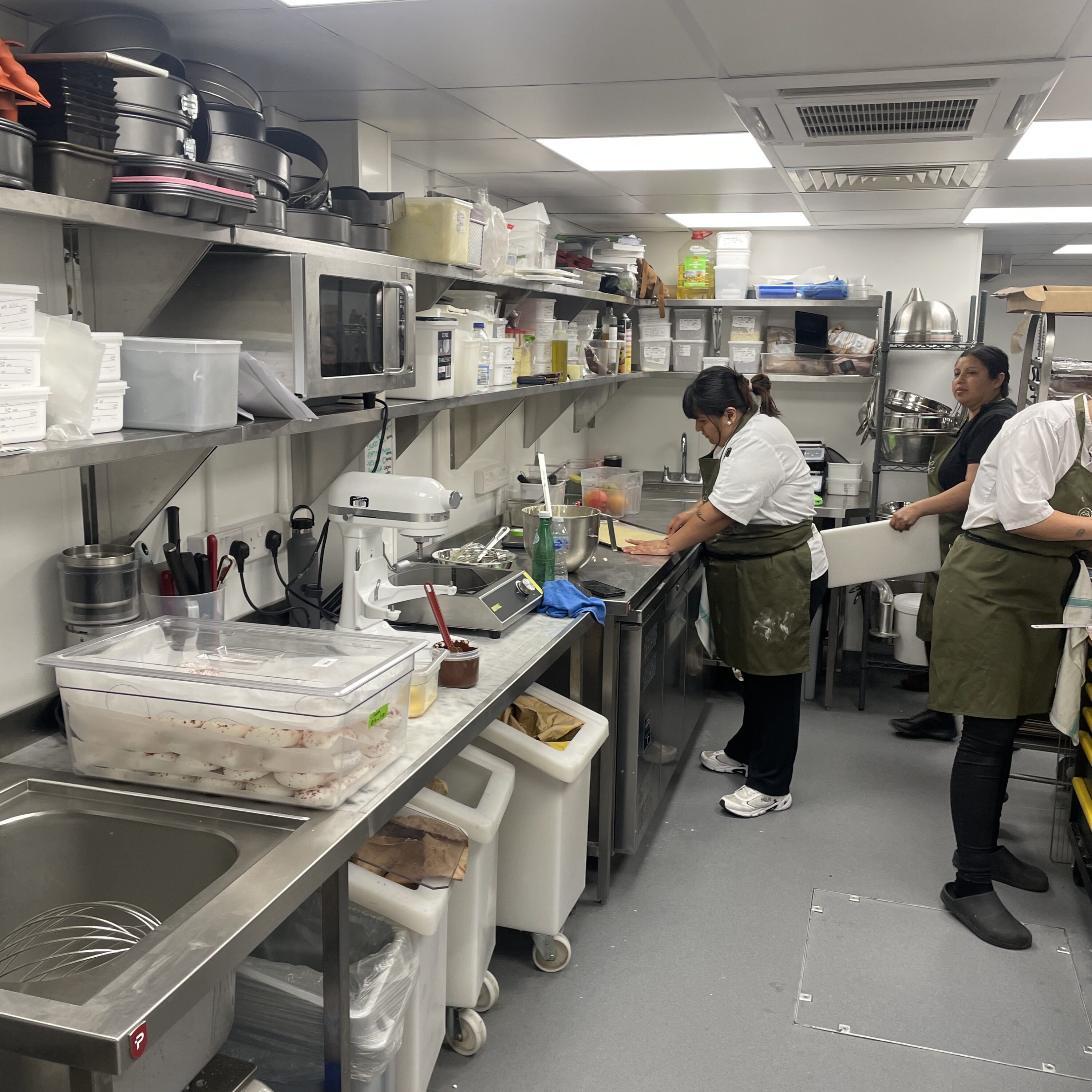 Busy kitchen with stainless steel surfaces, featuring chefs in white uniforms and green aprons preparing food.