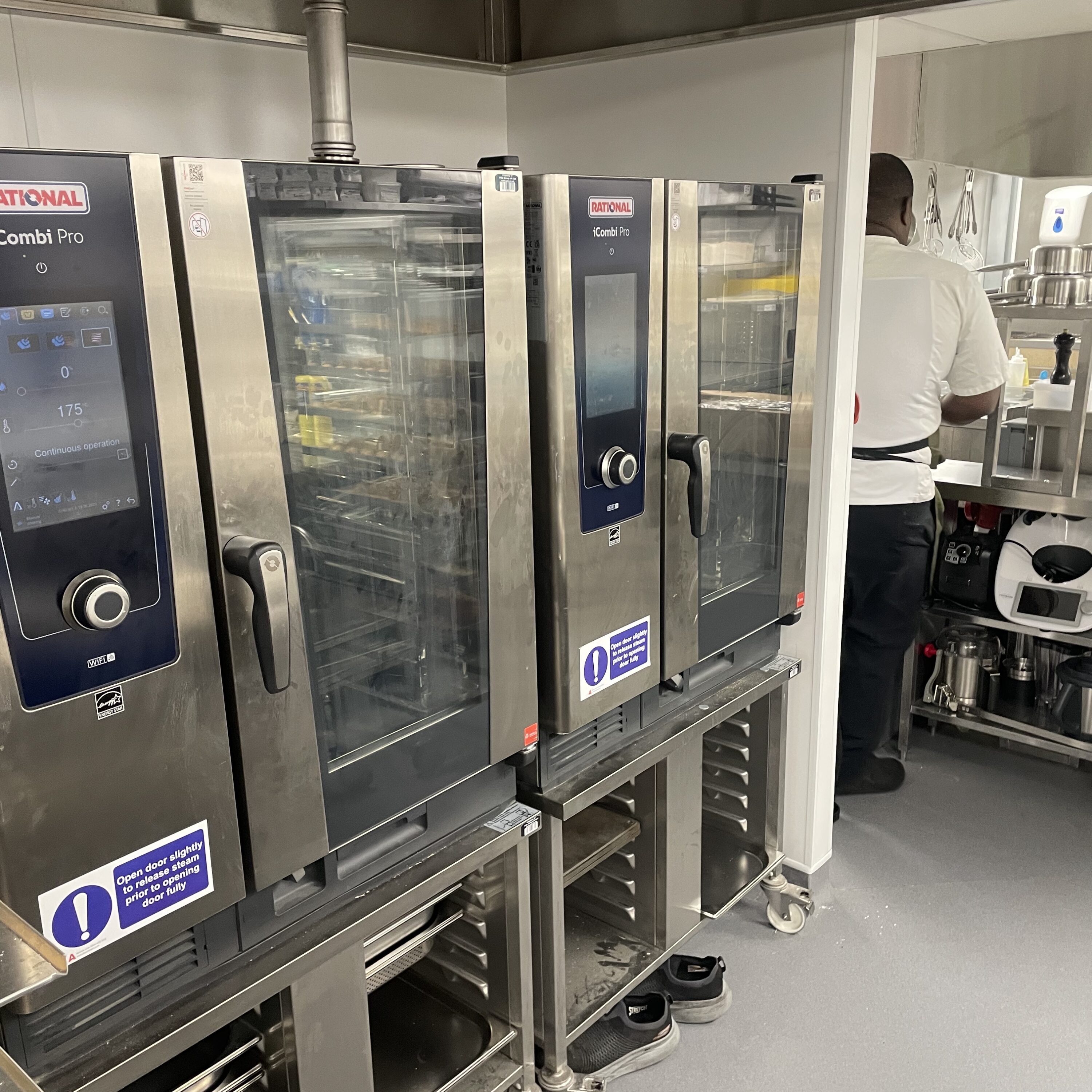 Stainless steel ovens line a commercial kitchen, with a chef visible in the background.