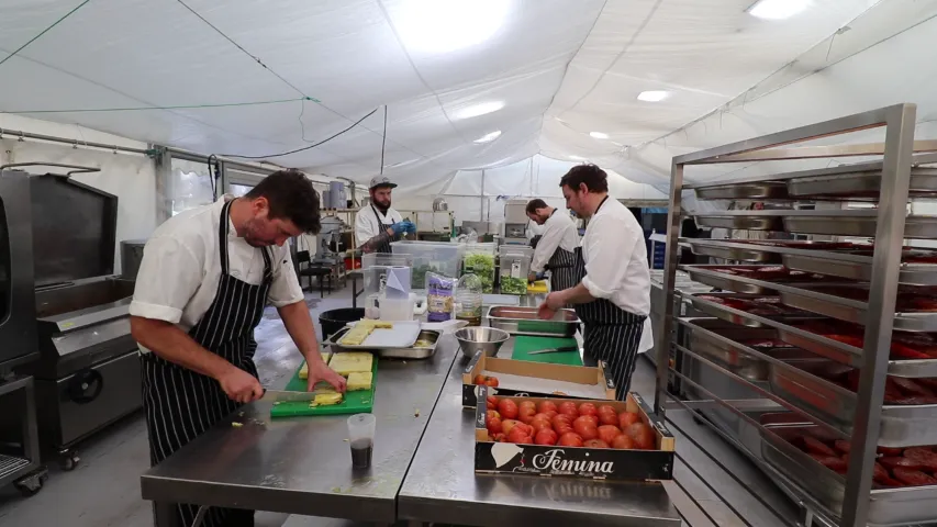 Chefs in a bright kitchen prepare ingredients on green chopping boards, with boxes of tomatoes visible.