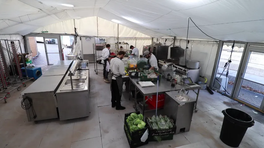 Busy kitchen with stainless steel equipment, chefs preparing food under a white tent.