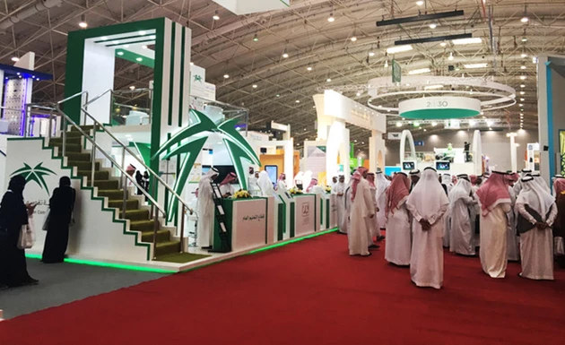 Exhibition hall featuring green and white displays, with attendees in traditional attire on a red carpet.