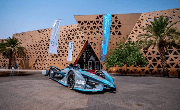 A sleek turquoise racing car is parked outside a modern building with geometric patterns.