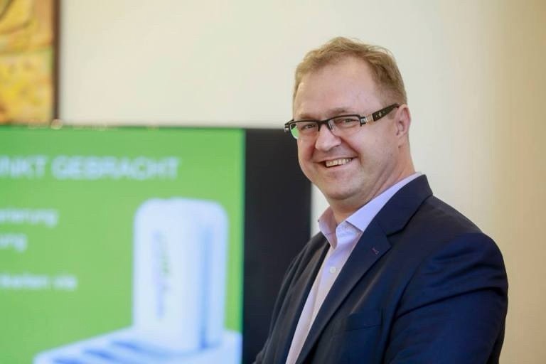 Smiling man in a dark suit with glasses, standing beside a green promotional display.