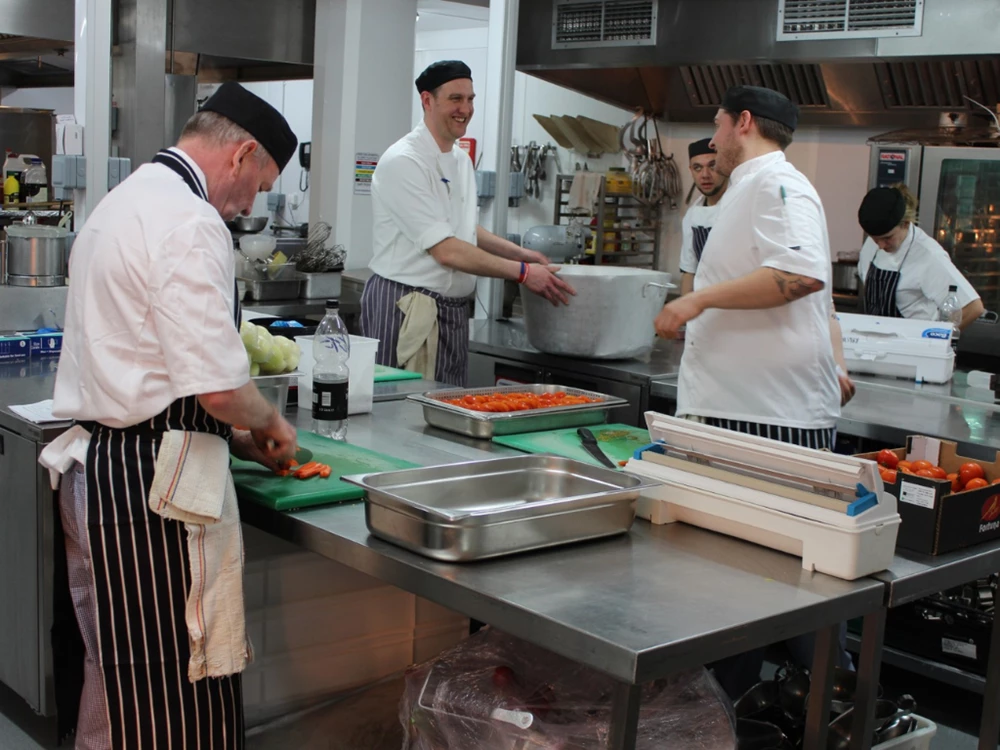 Chefs in white uniforms prepare food in a busy kitchen, featuring green cutting boards and stainless steel surfaces.
