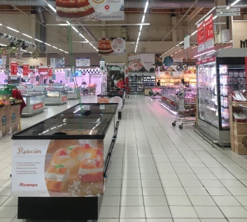 Brightly lit supermarket aisle featuring refrigerated displays and tiled flooring, with various products visible.