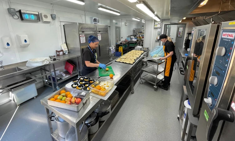 Two chefs prepare food in a stainless steel kitchen, with colourful fruits on the counter.