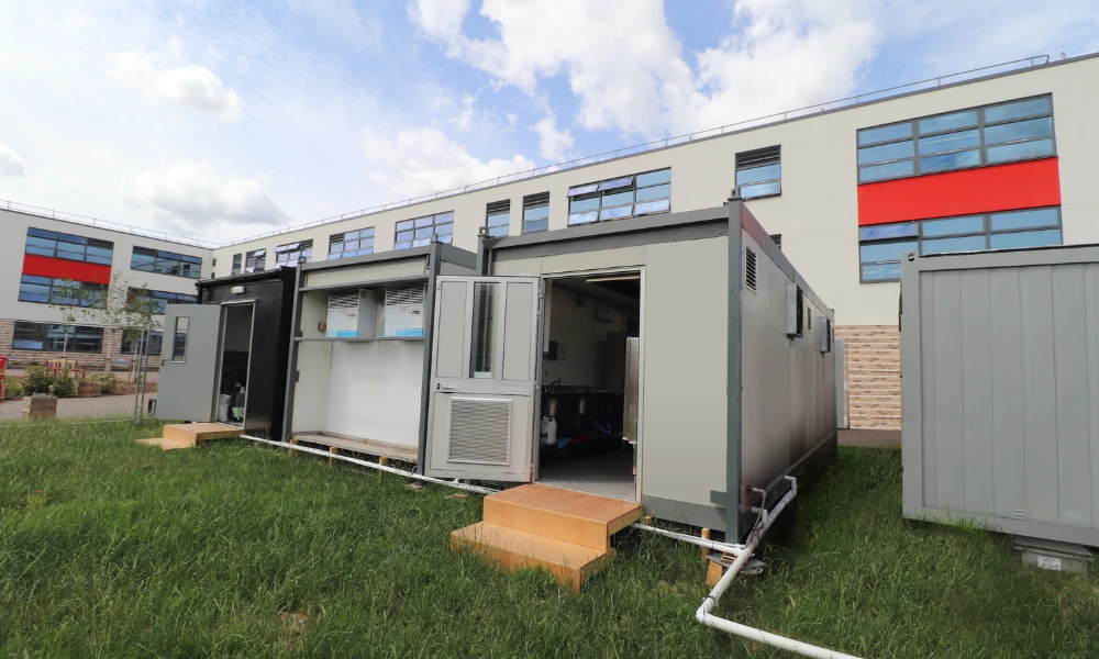Temporary kitchen unit in grey, with wooden steps, set against a building and blue sky.