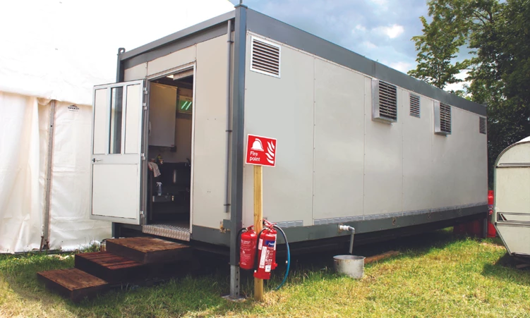 Temporary kitchen unit with a grey exterior, featuring an open door and fire safety equipment nearby.