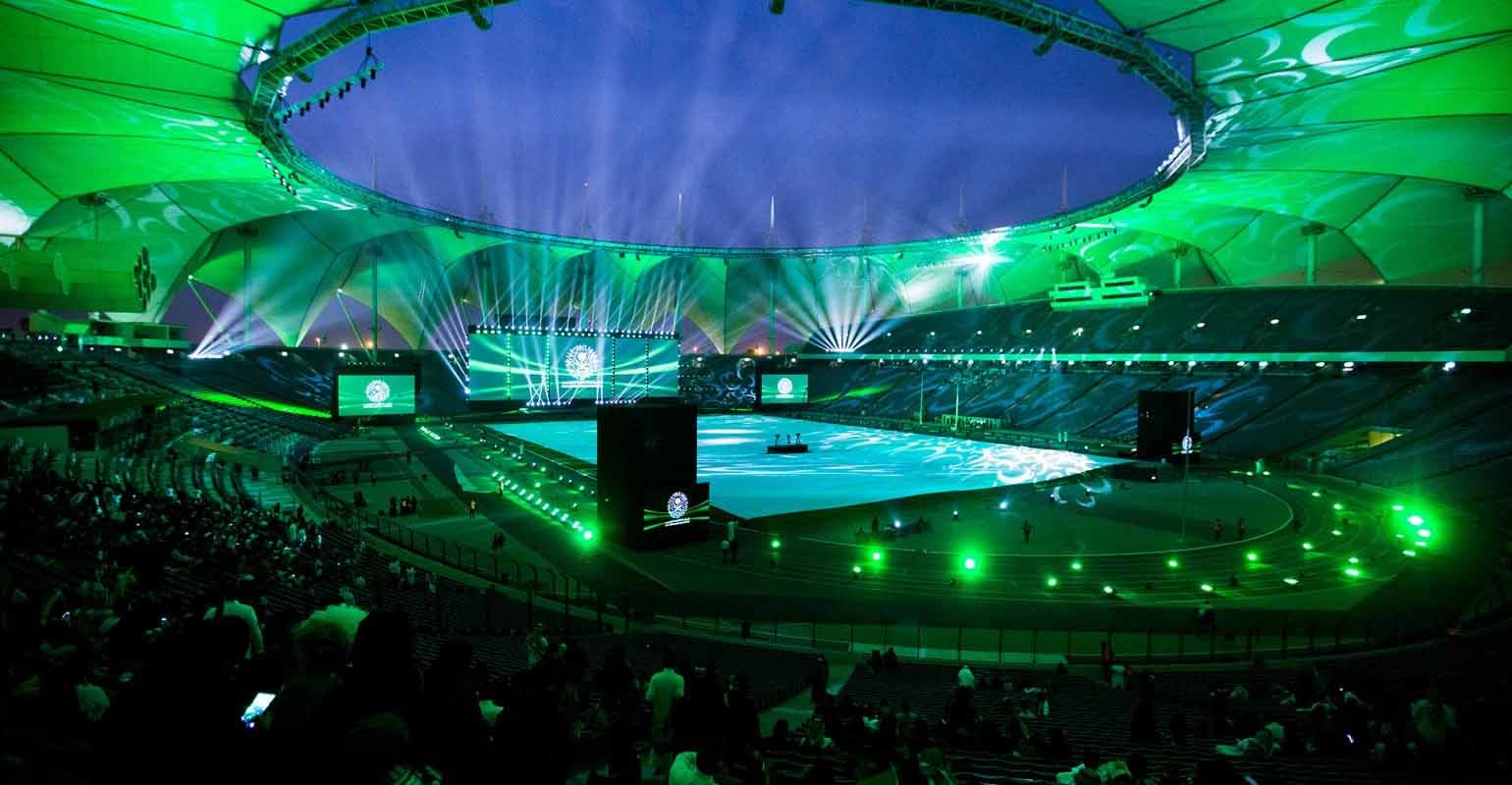 A vibrant green-lit stadium with a large stage and dramatic lighting effects during an event.
