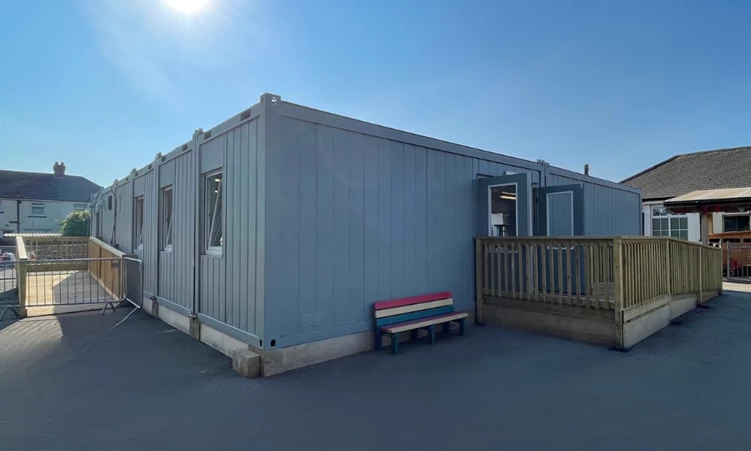 Light blue temporary building with a wooden deck and a red, white, and blue bench.