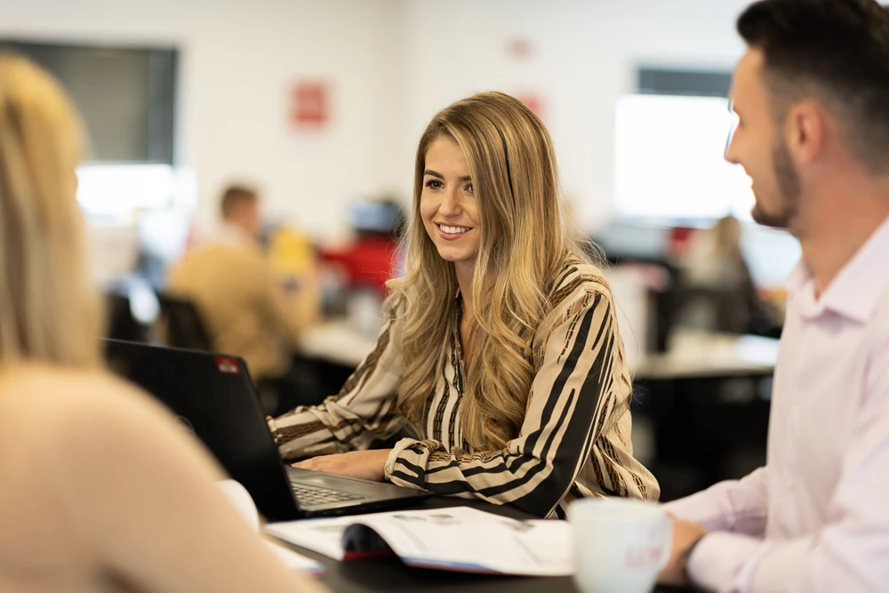 A woman with long, wavy blonde hair smiles while working on a laptop in a bright office.