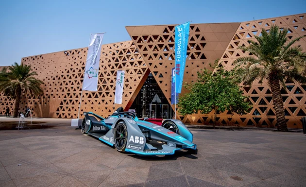 A turquoise racing car parked outside a modern building with a geometric facade and greenery.
