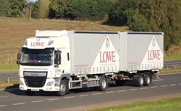 Lowe-branded articulated truck driving on a road, surrounded by green fields and trees.