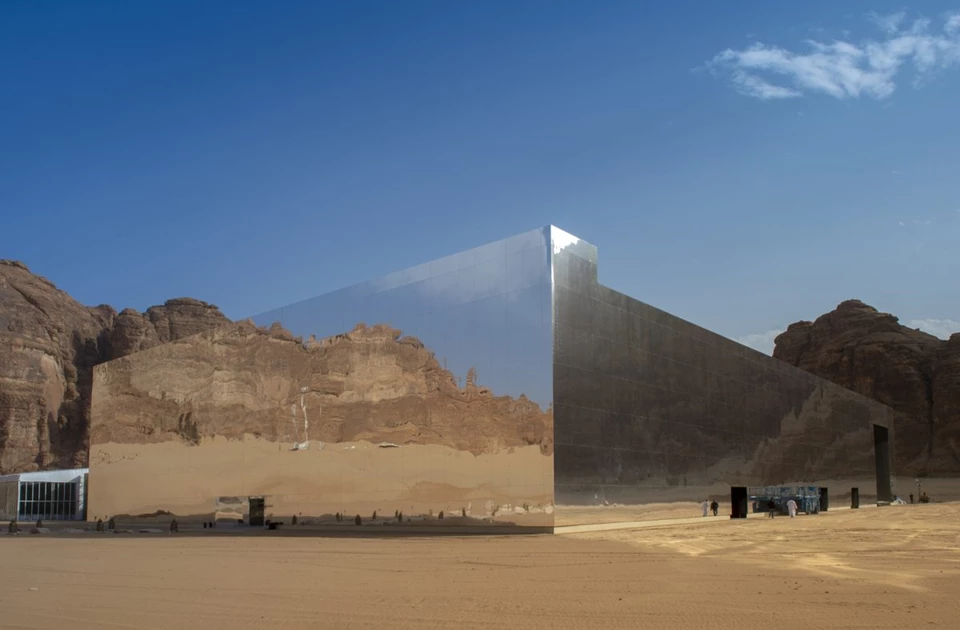 A large, reflective structure stands against a backdrop of sandy desert and rocky mountains.