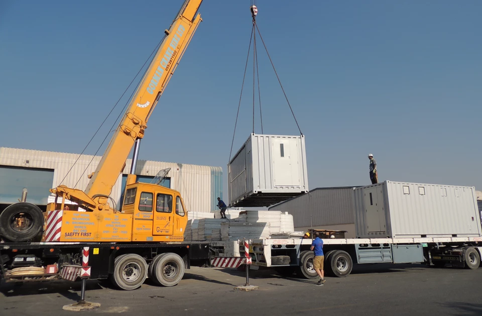 A yellow crane lifts a white container onto a truck in a clear, sunny environment.
