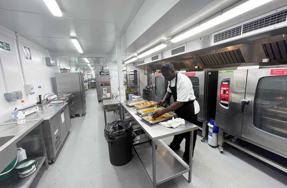 A chef in a white uniform prepares food on a stainless steel table in a temporary kitchen.