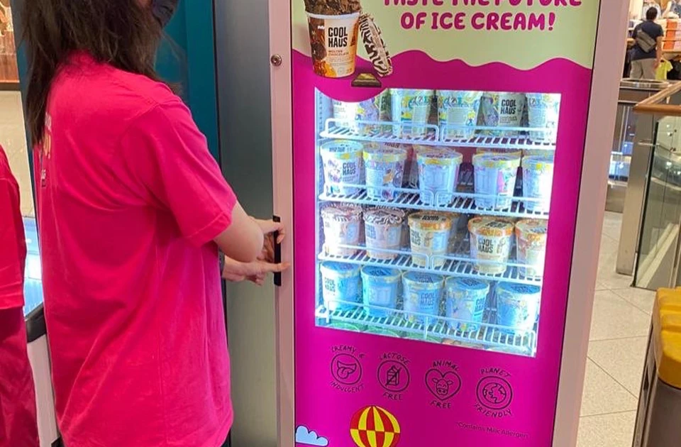 A person in a pink shirt operates an ice cream vending machine displaying various colourful tubs.