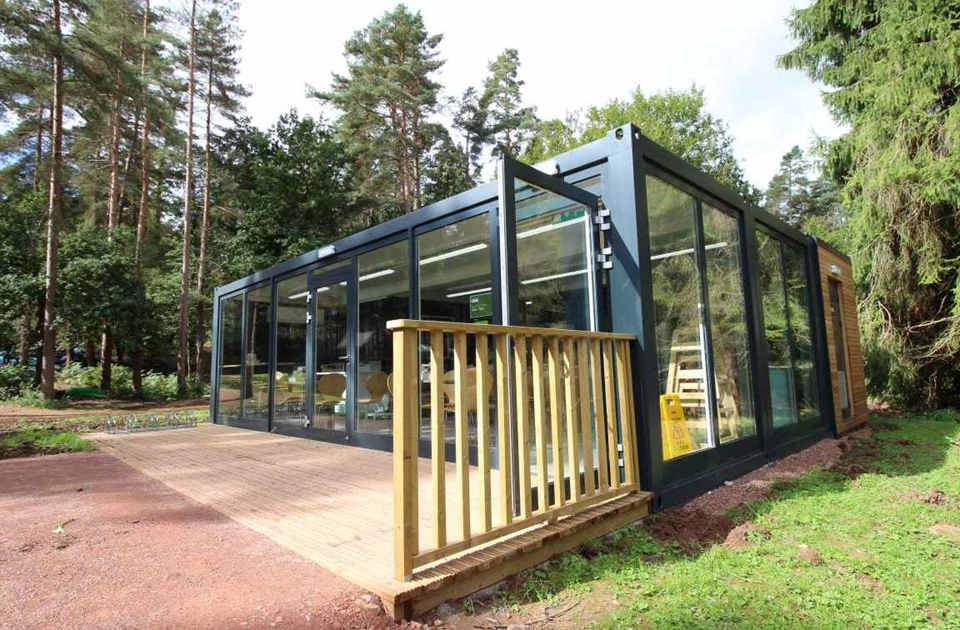 Modular café with large glass windows and a wooden deck, surrounded by trees in a natural setting.