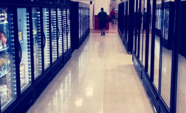 A person walks down a brightly lit aisle between glass-fronted refrigeration units.