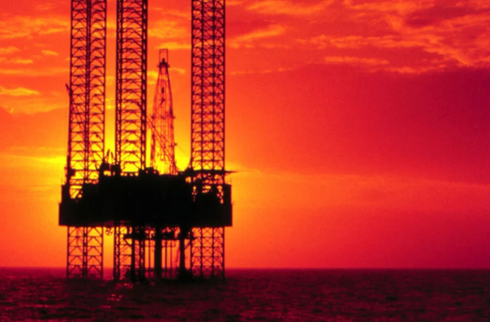 Oil rig silhouetted against a vibrant orange sunset over the ocean.