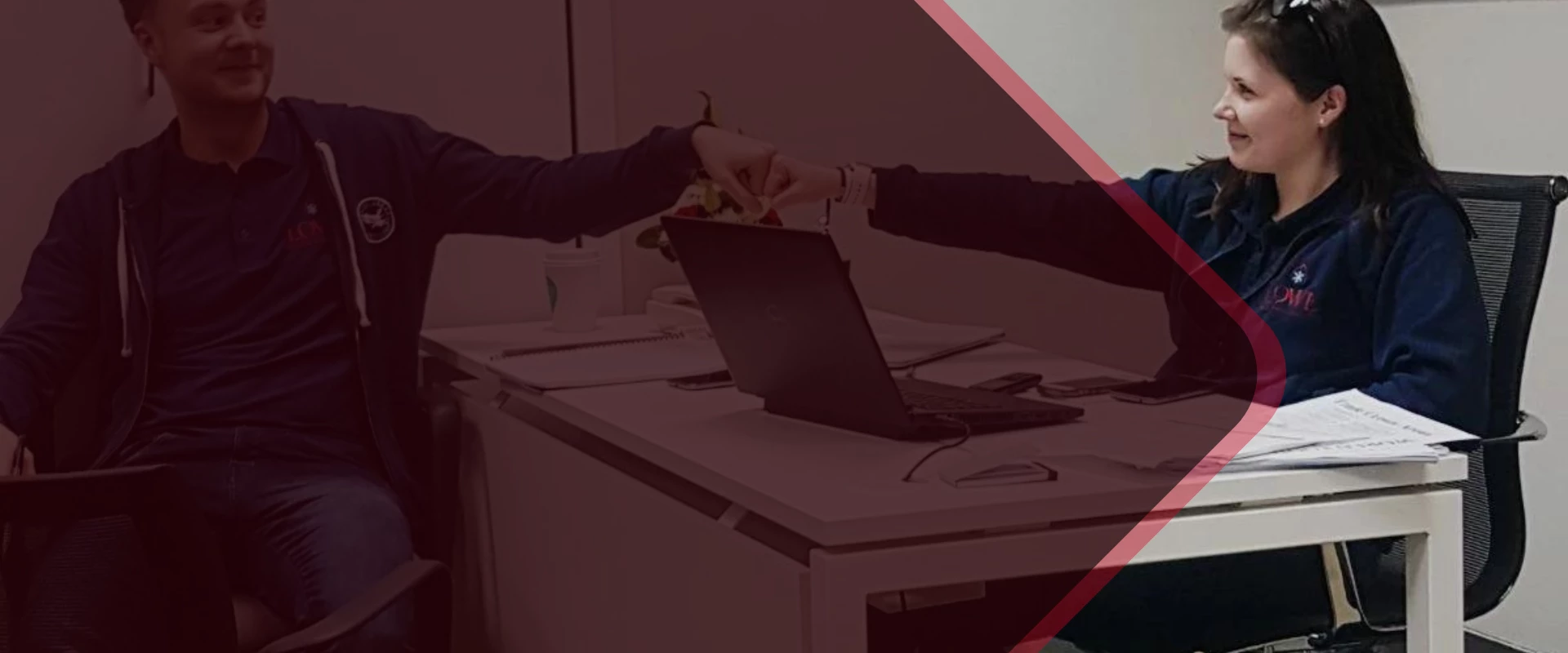Hands exchanging a document over a laptop on a white desk, with a dark red background.