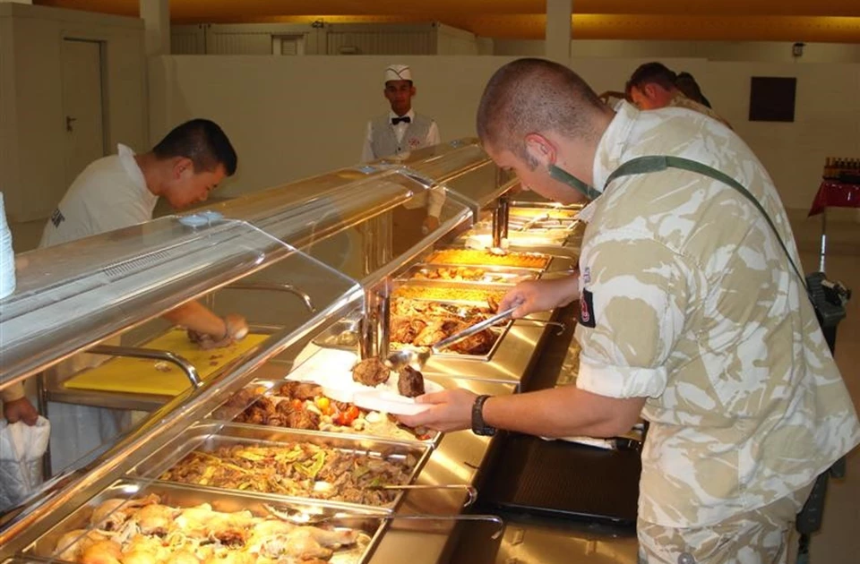 Soldiers in camouflage uniforms serve themselves from a buffet with various dishes in a temporary kitchen.