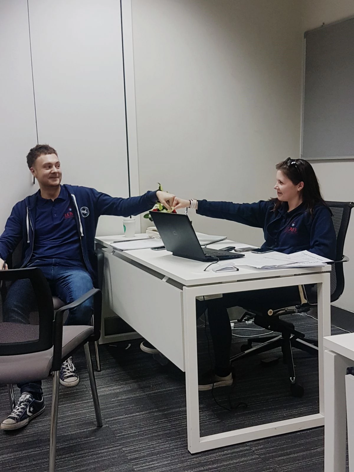 Two colleagues in navy blue hoodies fist bump across a white desk with a laptop.