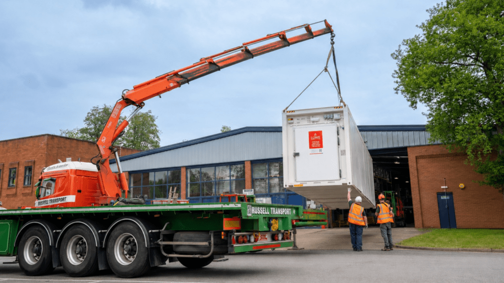 A crane lifts a white cold storage container from a green truck, with workers in orange vests nearby.