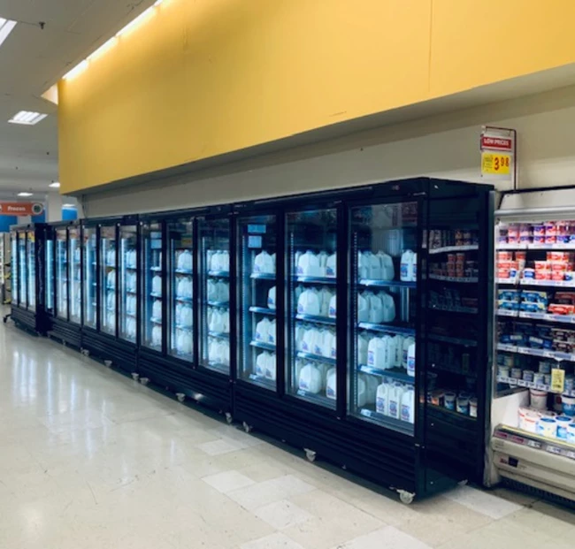 Rows of glass-fronted refrigerators displaying various products, set against a bright yellow wall.